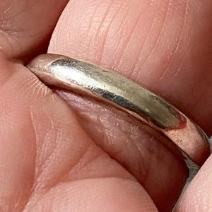 May include: A silver ring with a smooth, rounded band. The ring is held between fingers, showcasing its polished surface and simple design. The ring's metallic sheen reflects light, highlighting its minimalist style.