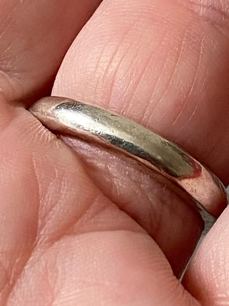 May include: A silver ring with a smooth, rounded band. The ring is held between fingers, showcasing its polished surface and simple design. The ring's metallic sheen reflects light, highlighting its minimalist style.