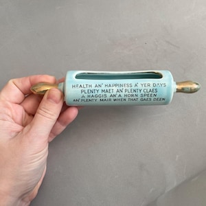 以下が含まれることがあります： 金色のハンドルが付いた、水色のセラミック製めん棒型の花瓶。 花瓶には長方形の開口部があり、「HEALTH AN' HAPPINESS A' YER DAYS...」というテキストが黒い文字で表示されています。 花瓶は人の手で持たれています。