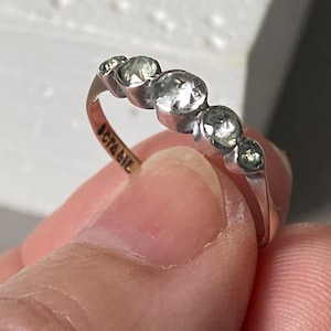 Op de afbeelding: Een zilverkleurige ring met vijf ronde, heldere, gefacetteerde stenen op een rij. De ringband is dun en van metaal, met de tekst "JCT&IE" duidelijk gegraveerd aan de binnenzijde, zichtbaar tegen de metalen afwerking van de ring.