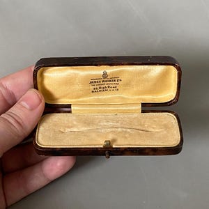 May include: An open antique jewelry box with a tortoiseshell pattern. The interior is lined with yellow velvet. The box is marked "JAMES WALKER LTD THE LONDON JEWELLERS 85 High Road BALHAM S.W.12".