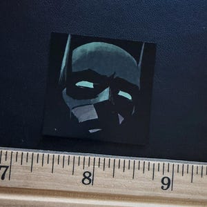 May include: A square sticker featuring a stylised illustration of a dark, masked character with pointed ears and glowing light blue eyes. The sticker is positioned above a wooden ruler showing measurements in centimetres, with the sticker width at 2.5 cm.