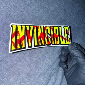 May include: A rectangular sticker featuring the word INVINCIBLE in bold, black-outlined capital letters. The interior of the letters displays a vibrant pattern of bright yellow and red flame-like shapes against a white background border.