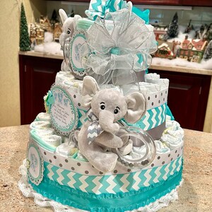 May include: A three-tiered diaper cake decorated with turquoise and gray chevron and polka dot patterned fabric. The top tier features a stuffed elephant holding a teething ring. The cake is topped with a turquoise and silver bow.