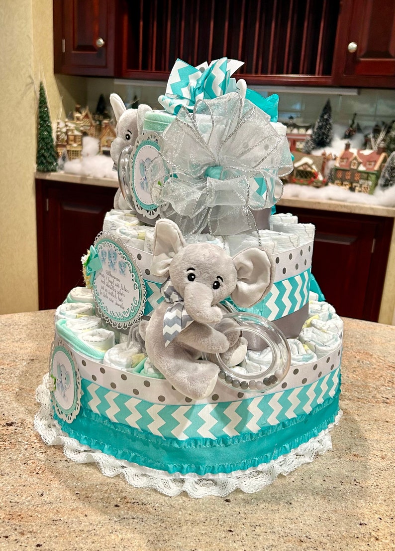 May include: A three-tiered diaper cake decorated with turquoise and gray chevron and polka dot patterned fabric. The top tier features a stuffed elephant holding a teething ring. The cake is topped with a turquoise and silver bow.