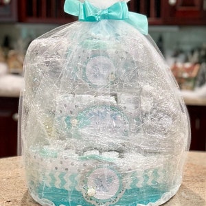 May include: A turquoise and white diaper cake with a white tulle and light blue ribbon bow. The cake is decorated with silver glitter and a snowflake design.