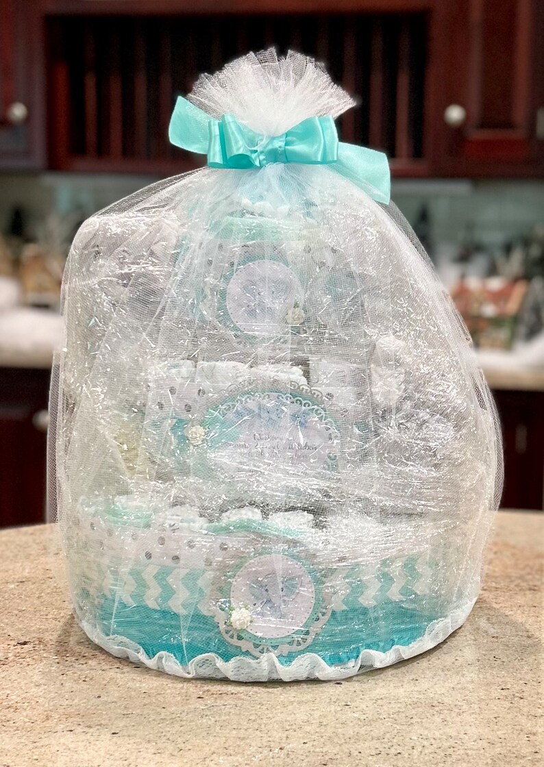 May include: A turquoise and white diaper cake with a white tulle and light blue ribbon bow. The cake is decorated with silver glitter and a snowflake design.