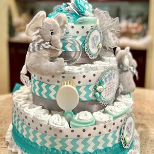 May include: A three-tiered diaper cake decorated with gray and teal ribbon, a gray elephant plush toy, and a white brush. The cake is decorated with a white lace trim and a small white flower.