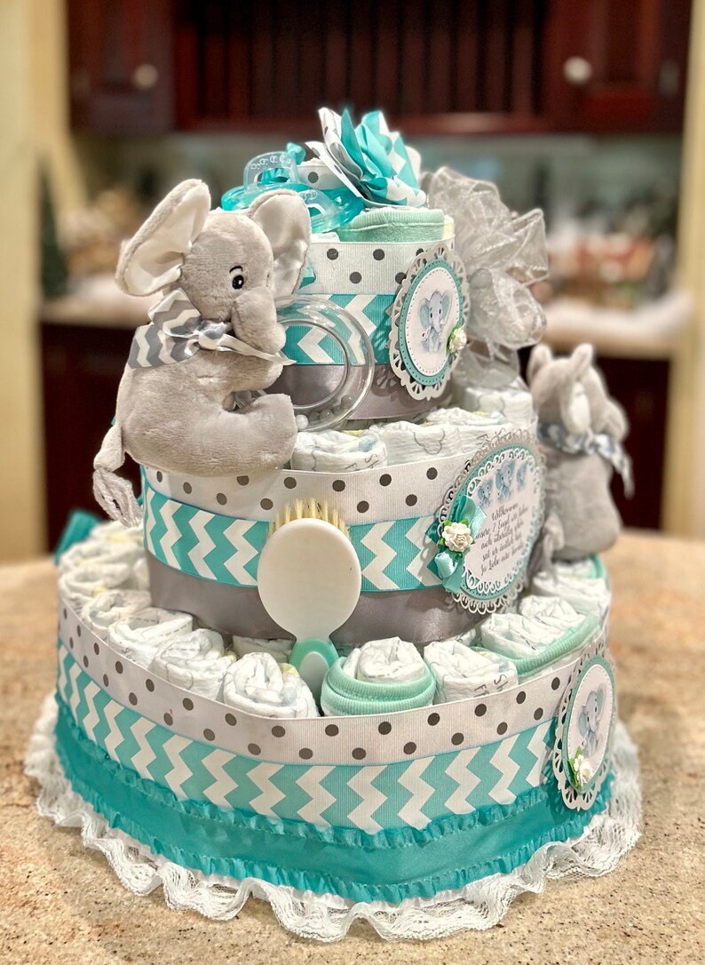 May include: A three-tiered diaper cake decorated with gray and teal ribbon, a gray elephant plush toy, and a white brush. The cake is decorated with a white lace trim and a small white flower.