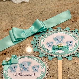 May include: Two teal blue and white paper cut-out circles with a decorative border. The circles feature a watercolor illustration of three baby elephants. One circle has the text "Willkommen!" and a teal blue bow. The other circle has a teal blue bow and a white flower.