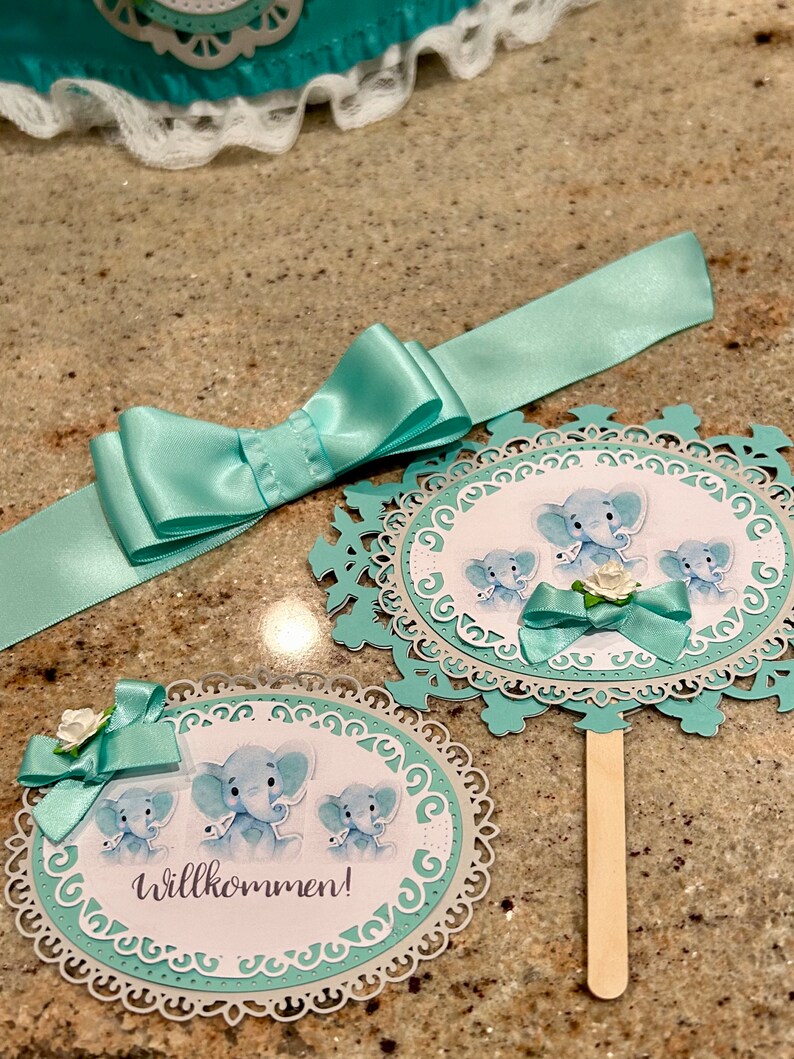 May include: Two teal blue and white paper cut-out circles with a decorative border. The circles feature a watercolor illustration of three baby elephants. One circle has the text "Willkommen!" and a teal blue bow. The other circle has a teal blue bow and a white flower.