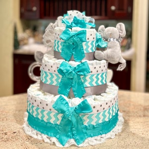 May include: A three-tiered diaper cake decorated with gray and turquoise chevron and polka dot patterned ribbon. The top tier features a gray plush elephant toy.
