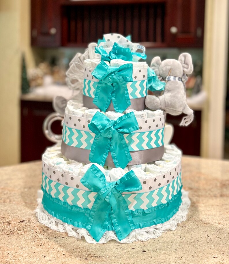 May include: A three-tiered diaper cake decorated with gray and turquoise chevron and polka dot patterned ribbon. The top tier features a gray plush elephant toy.