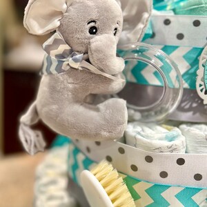 May include: A gray elephant plush toy with a gray and white bow tie is holding a clear plastic teething ring. The toy is on top of a diaper cake decorated with blue and gray chevron patterned ribbon, white polka dot ribbon, and a white brush.