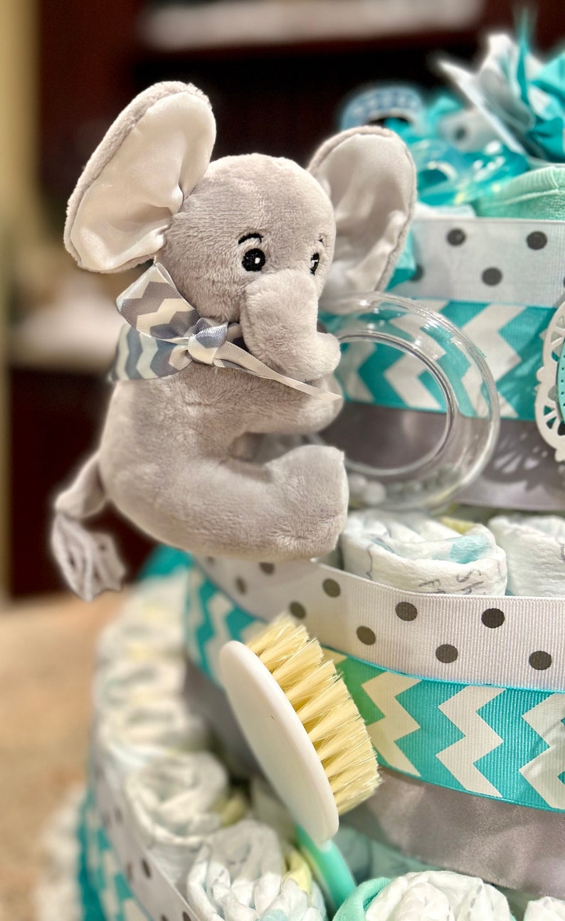 May include: A gray elephant plush toy with a gray and white bow tie is holding a clear plastic teething ring. The toy is on top of a diaper cake decorated with blue and gray chevron patterned ribbon, white polka dot ribbon, and a white brush.