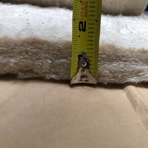 Op de afbeelding: Close-up van een dik, gebroken wit vezelmateriaal, mogelijk isolatie of vulling, met een gele meetlint die een dikte van ongeveer 5 cm aangeeft. De tape is gemarkeerd in voet en inch.