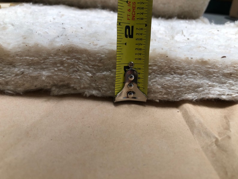 Op de afbeelding: Close-up van een dik, gebroken wit vezelmateriaal, mogelijk isolatie of vulling, met een gele meetlint die een dikte van ongeveer 5 cm aangeeft. De tape is gemarkeerd in voet en inch.