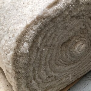 Op de afbeelding: Een grote rol van off-white, getextureerde katoenen watten. De watten zijn strak opgerold, waardoor de gelaagde textuur van het materiaal zichtbaar wordt. Kleine donkere vlekken zijn zichtbaar in het katoen. De rol ligt op een bruin papieren vel.