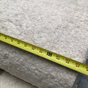Op de afbeelding: Een opgerold, cr&egrave;mekleurig, vezelig materiaal met een gele meetlint eroverheen. De tape toont metingen tot 68,5 cm. Het materiaal lijkt een natuurvezelproduct te zijn, mogelijk voor isolatie of vulling.