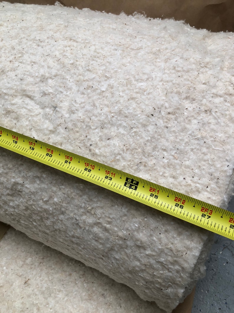 Op de afbeelding: Een opgerold, cr&egrave;mekleurig, vezelig materiaal met een gele meetlint eroverheen. De tape toont metingen tot 68,5 cm. Het materiaal lijkt een natuurvezelproduct te zijn, mogelijk voor isolatie of vulling.