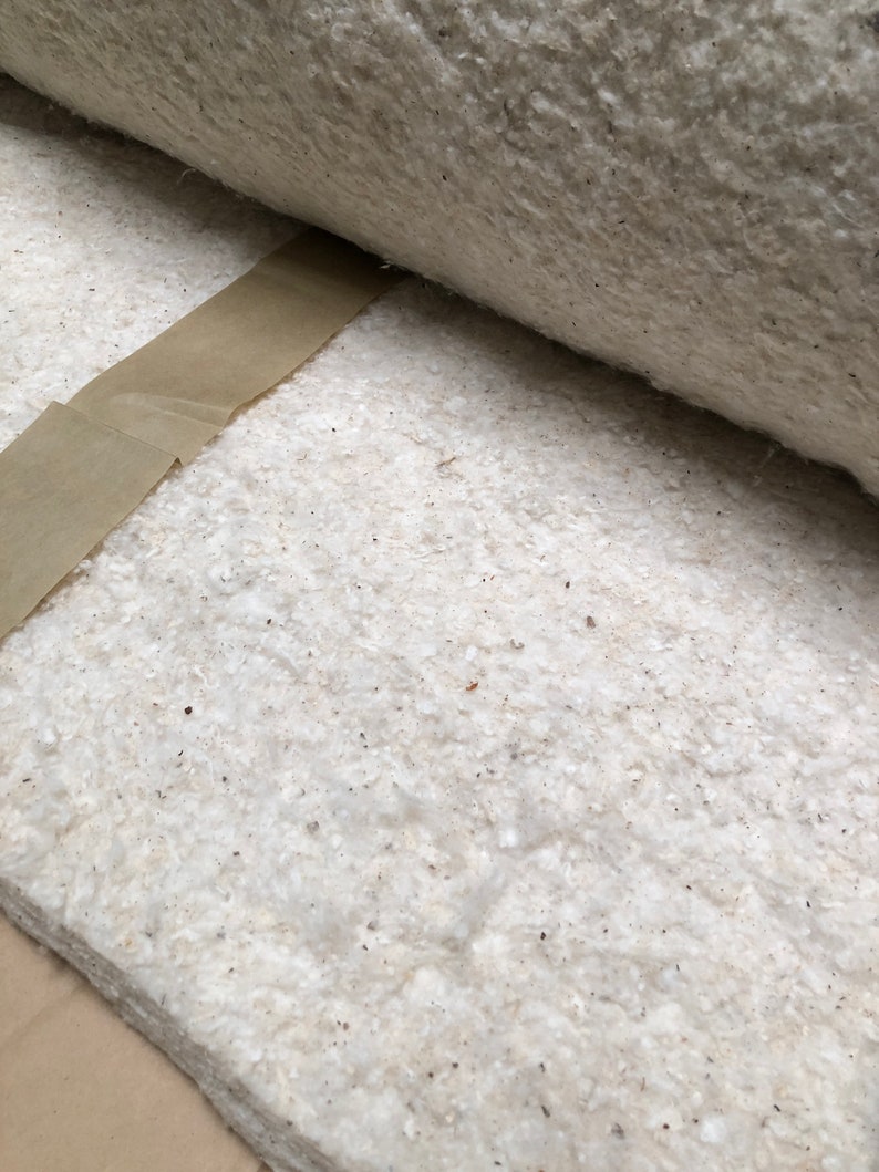 Op de afbeelding: Close-up van een opgerold, gebroken wit, getextureerd materiaal met een bruine papieren strip. Het materiaal lijkt een dikke, vezelige stof te zijn, mogelijk isolatie of vulling. De textuur is bespikkeld met kleine, donkere vlekjes.