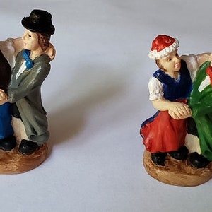 May include: Four small figurines of dancing couples. Each figurine features a man in a suit and hat, embracing a woman in a dress and bonnet. The couples are painted in various colors, including green, blue, red, and purple. They are standing on a brown base.