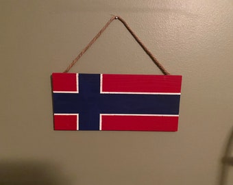 May include: A wooden sign painted with the Norwegian flag. The flag is red, white, and blue. The sign is hanging from a string.