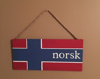 May include: A wooden sign with the Norwegian flag painted on it. The sign is hanging from a piece of twine and has the word "norsk" written in white letters on the blue portion of the flag.