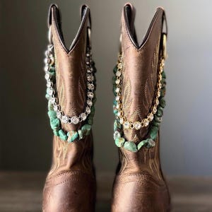 Könnte beinhalten: Ein Paar braune Leder-Cowboystiefel mit dekorativen Ketten, die um den Schaft drapiert sind. Jeder Stiefel ist mit einer Reihe klarer Strasssteine und einer Reihe rauer, türkisfarbener Steine verziert, was einen geschichteten Effekt erzeugt.