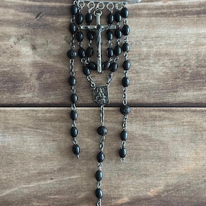 May include: A black rosary with oval beads and a silver crucifix pendant. The rosary is attached to a large safety pin. The background is a wooden surface.