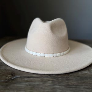 May include: A beige fedora hat with a wide brim and a pinched crown. The hat features a decorative band around the base of the crown, composed of small, rectangular, white, smooth-surfaced beads arranged in a continuous horizontal row.