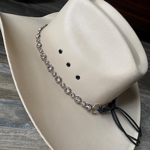 Pu&ograve; includere: Un cappello da cowboy color crema con finitura testurizzata, caratterizzato da una fascia di strass rotondi e trasparenti incastonati in argento attorno alla corona. Il cappello presenta tre occhielli di ventilazione neri e un laccio in pelle.