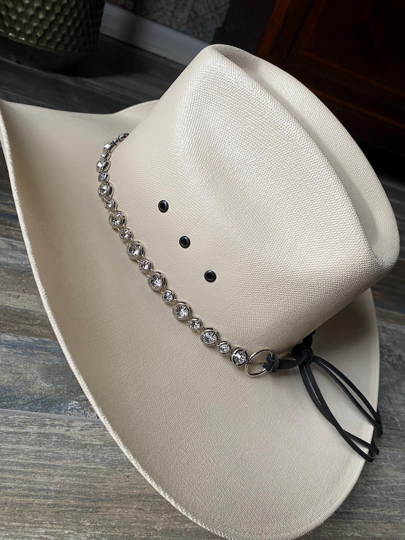 Pu&ograve; includere: Un cappello da cowboy color crema con finitura testurizzata, caratterizzato da una fascia di strass rotondi e trasparenti incastonati in argento attorno alla corona. Il cappello presenta tre occhielli di ventilazione neri e un laccio in pelle.