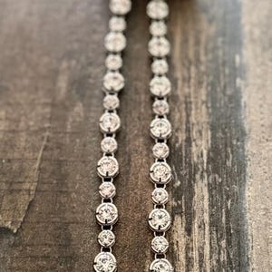 Pu&ograve; includere: Una collana tennis color argento caratterizzata da una fila continua di pietre rotonde, trasparenti e sfaccettate, incastonate in montature di metallo. La collana termina in alto con un nastro di tessuto nero annodato a cappio.