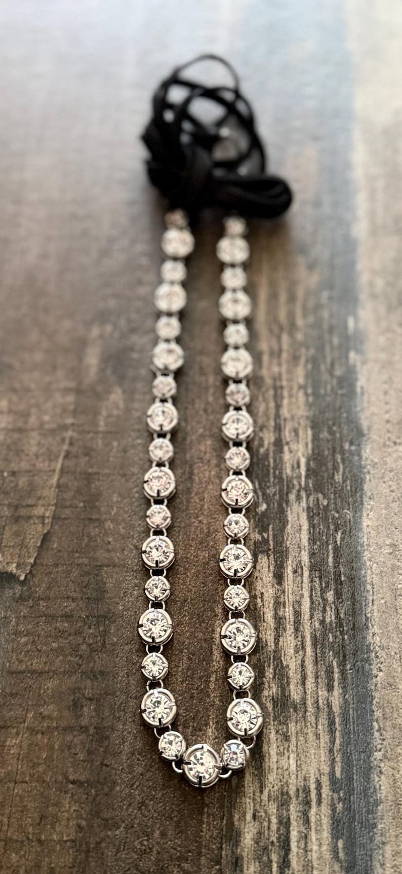 Pu&ograve; includere: Una collana tennis color argento caratterizzata da una fila continua di pietre rotonde, trasparenti e sfaccettate, incastonate in montature di metallo. La collana termina in alto con un nastro di tessuto nero annodato a cappio.