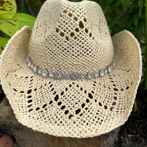 Pu&ograve; includere: Un cappello da cowboy in paglia intrecciata color beige chiaro con tesa larga e corona alta. Il cappello &egrave; decorato con una fascia di strass rotondi, trasparenti e sfaccettati, incastonati in metallo argentato attorno alla base della corona.