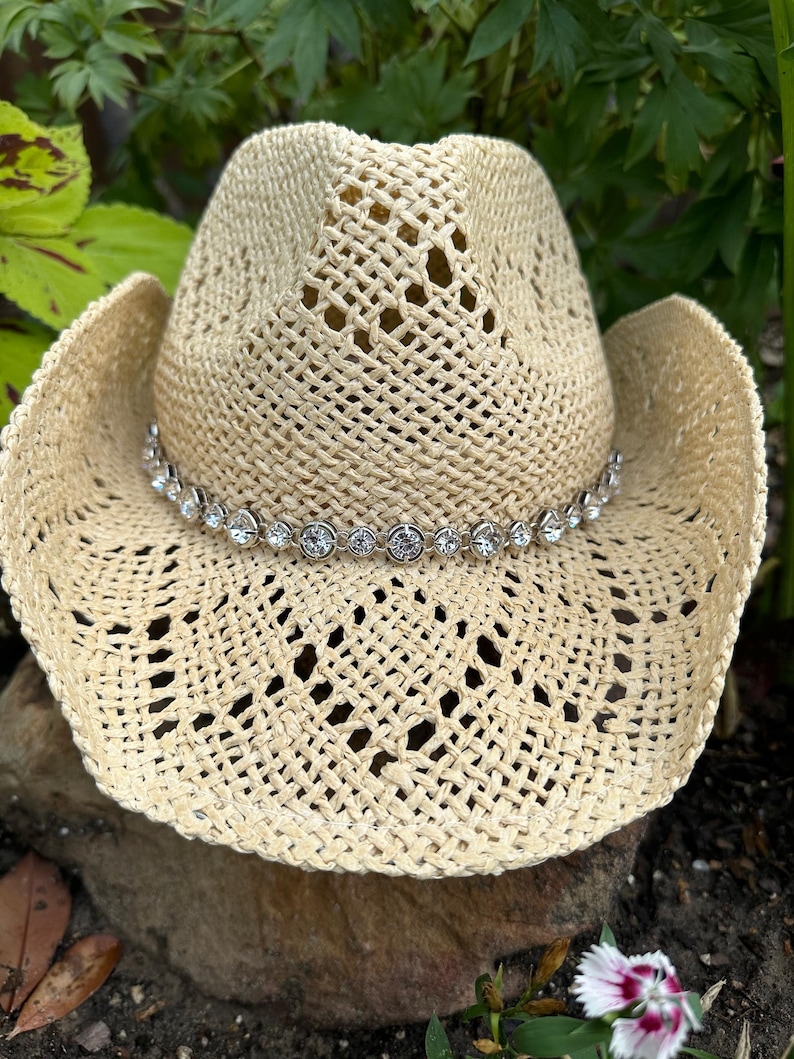 Pu&ograve; includere: Un cappello da cowboy in paglia intrecciata color beige chiaro con tesa larga e corona alta. Il cappello &egrave; decorato con una fascia di strass rotondi, trasparenti e sfaccettati, incastonati in metallo argentato attorno alla base della corona.