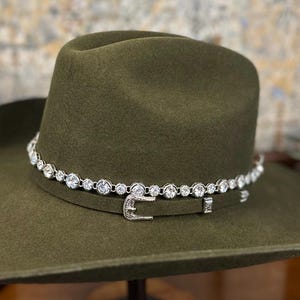 Pu&ograve; includere: Un cappello da cowboy in feltro verde oliva caratterizzato da una fascia argentata con chiusura a fibbia e una fila di strass rotondi, trasparenti e sfaccettati che circondano la corona sopra la fascia, su uno sfondo neutro e sfocato.