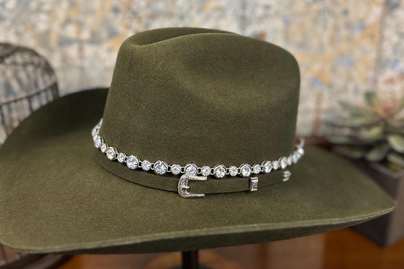 Pu&ograve; includere: Un cappello da cowboy in feltro verde oliva caratterizzato da una fascia argentata con chiusura a fibbia e una fila di strass rotondi, trasparenti e sfaccettati che circondano la corona sopra la fascia, su uno sfondo neutro e sfocato.