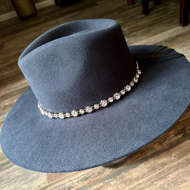 Pu&ograve; includere: Un cappello fedora blu scuro a tesa larga con corona strutturata e tesa piatta. Una singola fila di strass trasparenti, rotondi e sfaccettati, incastonati in una catena di metallo color argento, circonda la base della corona come fascia decorativa.