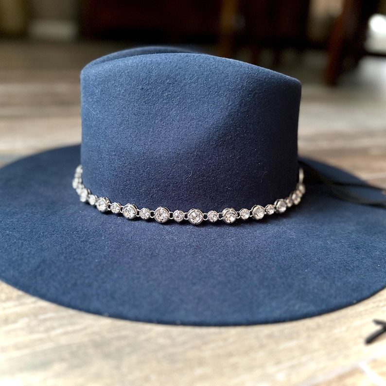 Pu&ograve; includere: Un cappello fedora blu navy a tesa larga con corona strutturata e tesa piatta. La base della corona &egrave; circondata da una catena color argento impreziosita da strass trasparenti, rotondi e sfaccettati che catturano la luce.