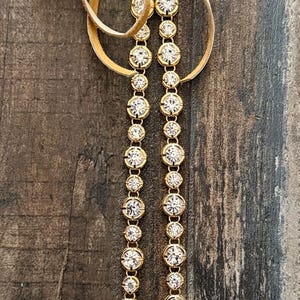 Pu&ograve; includere: Una collana composta da due fili paralleli di cristalli rotondi, trasparenti e sfaccettati, incastonati in montature di metallo dorato. I fili sono collegati in alto a un cordino in camoscio color beige chiaro annodato al centro.