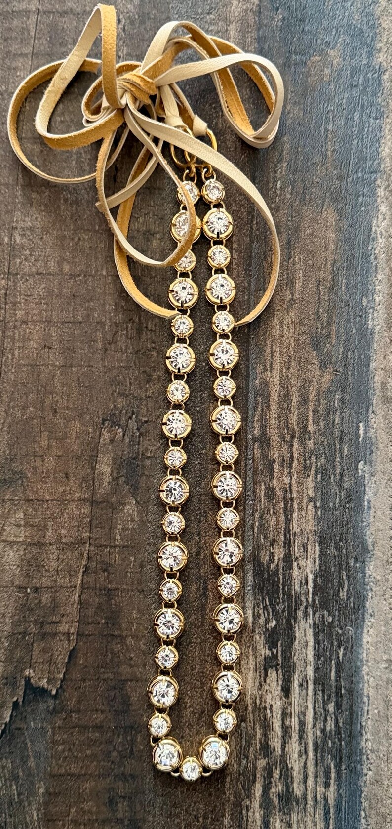 Pu&ograve; includere: Una collana composta da due fili paralleli di cristalli rotondi, trasparenti e sfaccettati, incastonati in montature di metallo dorato. I fili sono collegati in alto a un cordino in camoscio color beige chiaro annodato al centro.