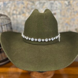 Pu&ograve; includere: Un cappello da cowboy in feltro verde oliva con una fascia color argento decorata da una fila continua di cristalli chiari, rotondi e sfaccettati che circondano la corona. Il cappello presenta una corona strutturata e una tesa larga.