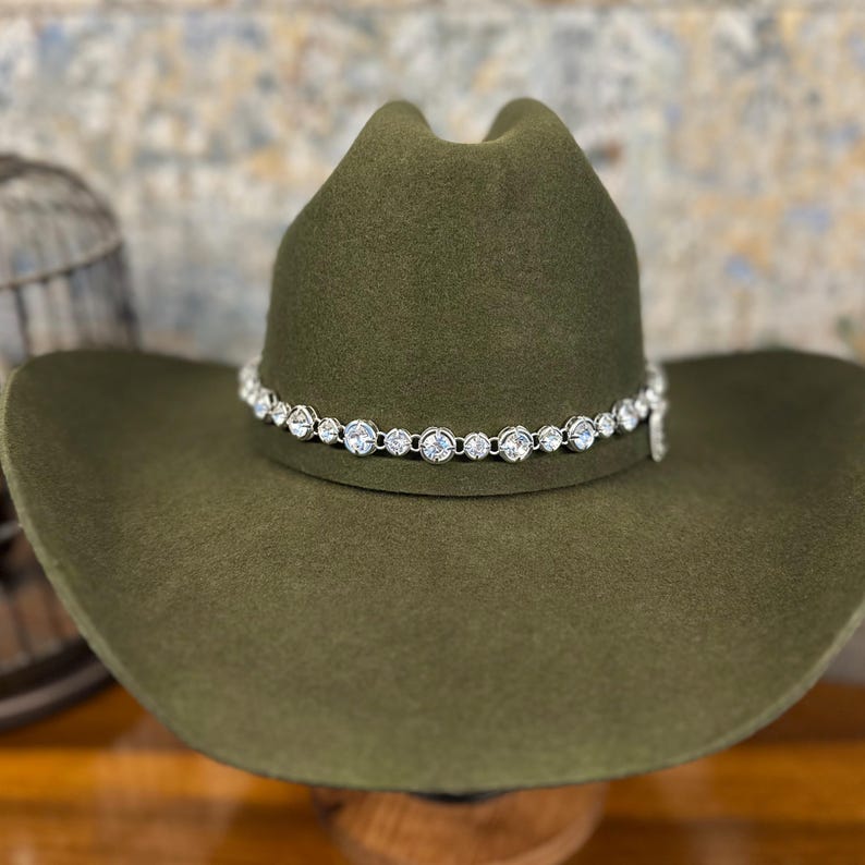 Pu&ograve; includere: Un cappello da cowboy in feltro verde oliva con una fascia color argento decorata da una fila continua di cristalli chiari, rotondi e sfaccettati che circondano la corona. Il cappello presenta una corona strutturata e una tesa larga.