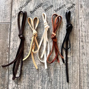Pu&ograve; includere: Cinque cordini in pelle annodati a cappio, etichettati Chocolate, Buckskin, White, Tobacco e Black. Ogni cordino presenta un colore distinto e un design a cappio annodato, mostrando la variet&agrave; di tonalit&agrave; disponibili per queste strisce.