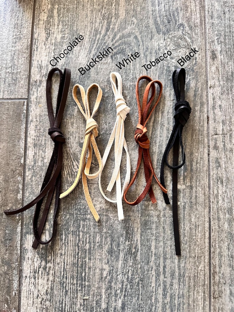 Pu&ograve; includere: Cinque cordini in pelle annodati a cappio, etichettati Chocolate, Buckskin, White, Tobacco e Black. Ogni cordino presenta un colore distinto e un design a cappio annodato, mostrando la variet&agrave; di tonalit&agrave; disponibili per queste strisce.
