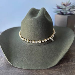 Pu&ograve; includere: Un cappello da cowboy in feltro verde oliva con tesa larga e corona pizzicata. Una fascia a catena color oro circonda la base della corona, decorata con una fila continua di strass rotondi, trasparenti e sfaccettati in montature dorate.