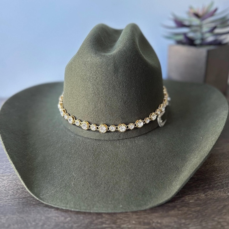 Pu&ograve; includere: Un cappello da cowboy in feltro verde oliva con tesa larga e corona pizzicata. Una fascia a catena color oro circonda la base della corona, decorata con una fila continua di strass rotondi, trasparenti e sfaccettati in montature dorate.