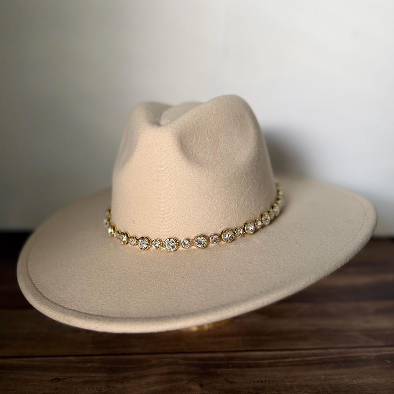 Pu&ograve; includere: Un cappello fedora in feltro beige con tesa larga e corona pizzicata. La base della corona &egrave; circondata da una fascia decorativa composta da piccoli cristalli rotondi e trasparenti sfaccettati, incastonati in una catena di metallo dorato.